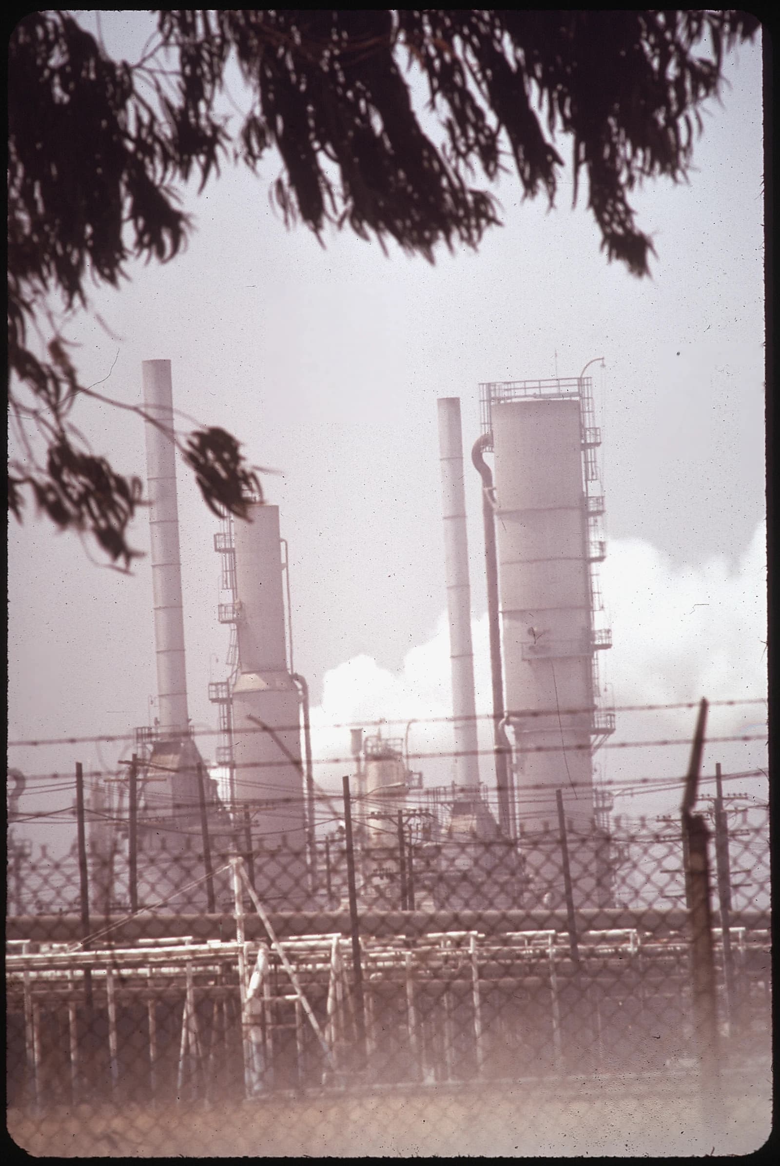 Mobil Oil refinery industrial structures seen through smog and a chain-link fence.