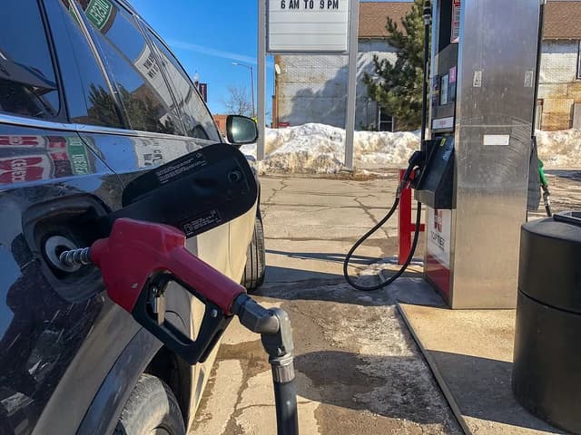 A red fuel nozzle inserted into the gas tank of a dark colored car at a gas station.