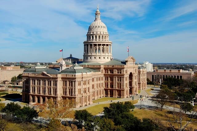 The Texas State Capitol building under a blue sky, surrounded by grounds and trees.