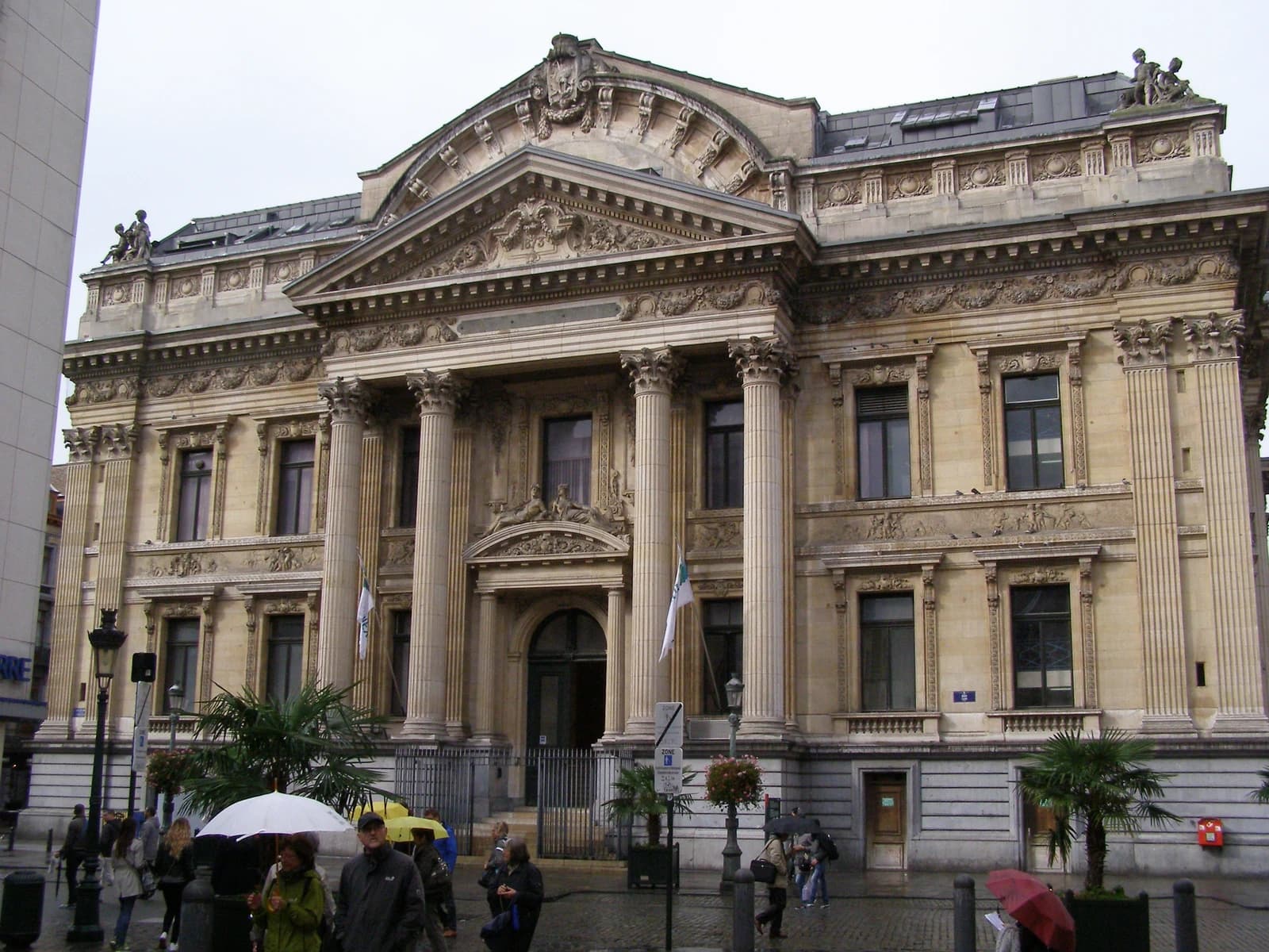 People walk with umbrellas in front of the Brussels Stock Exchange building on a cloudy day.