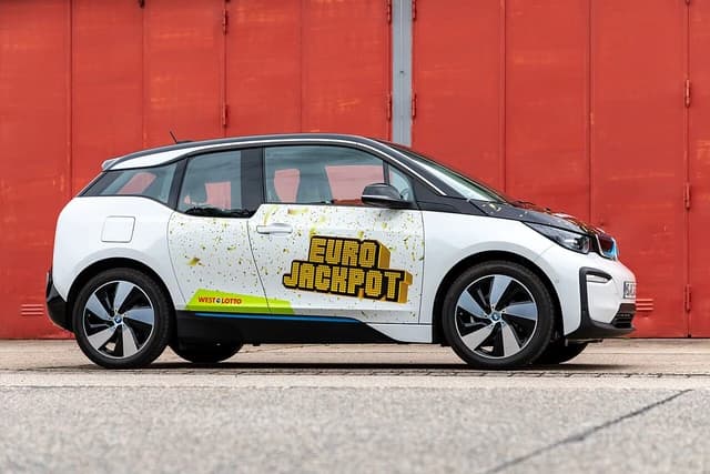 White BMW i3 electric car with promotional Euro Jackpot livery parked next to a red building.