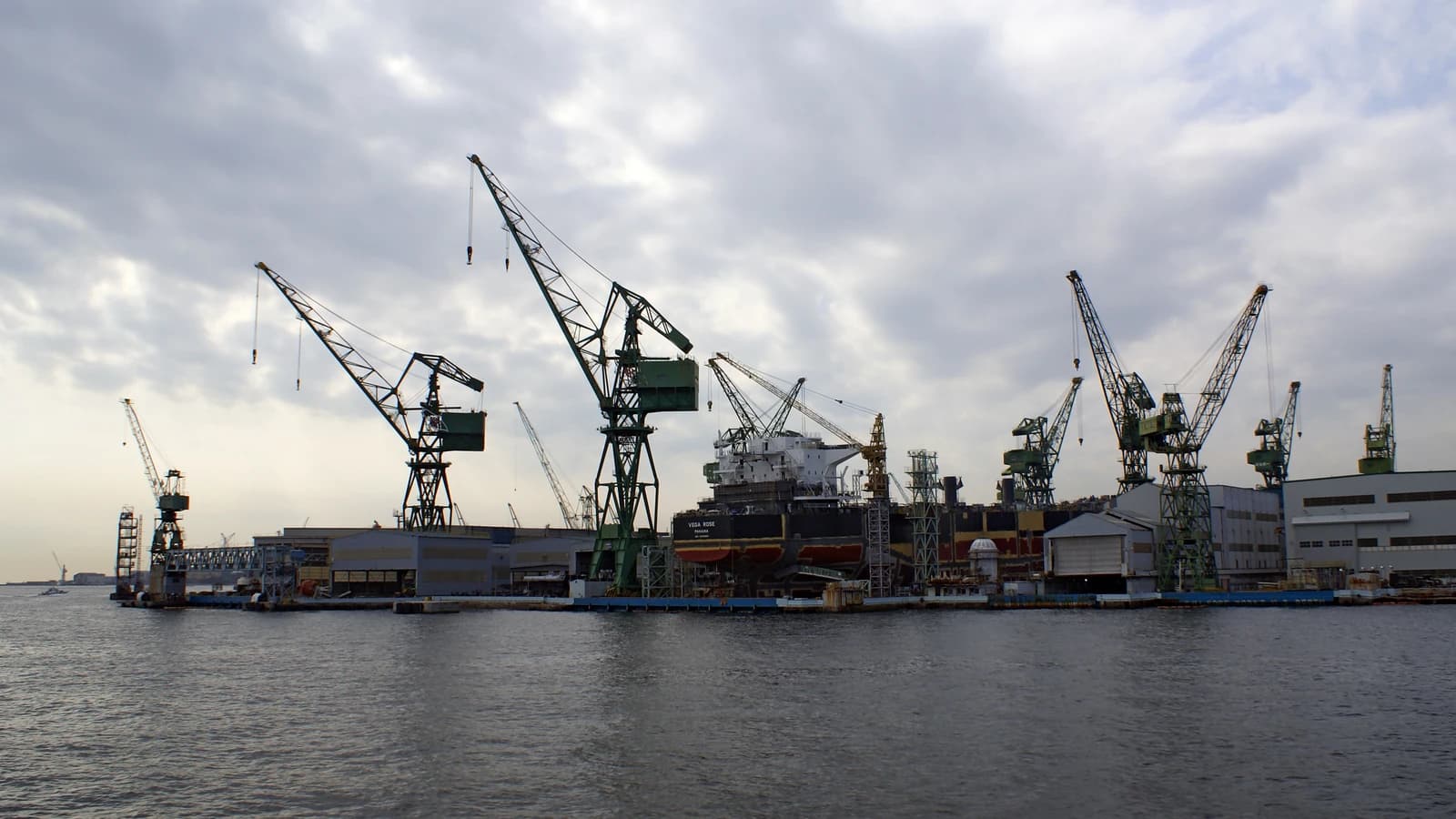 A large shipyard with multiple cranes and a ship in a dry dock, under a cloudy sky.