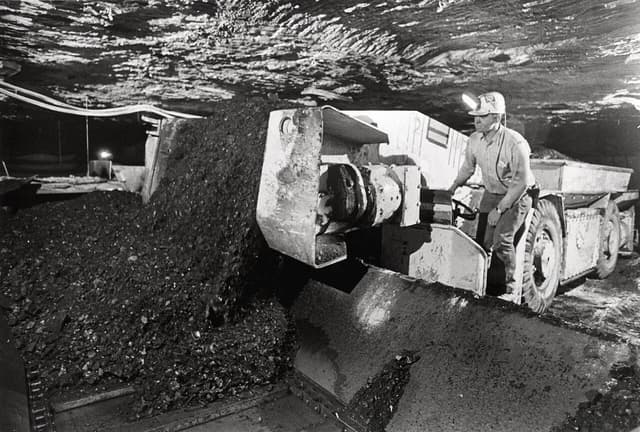 Coal miner operating large mining equipment underground, loading raw coal onto a conveyor.