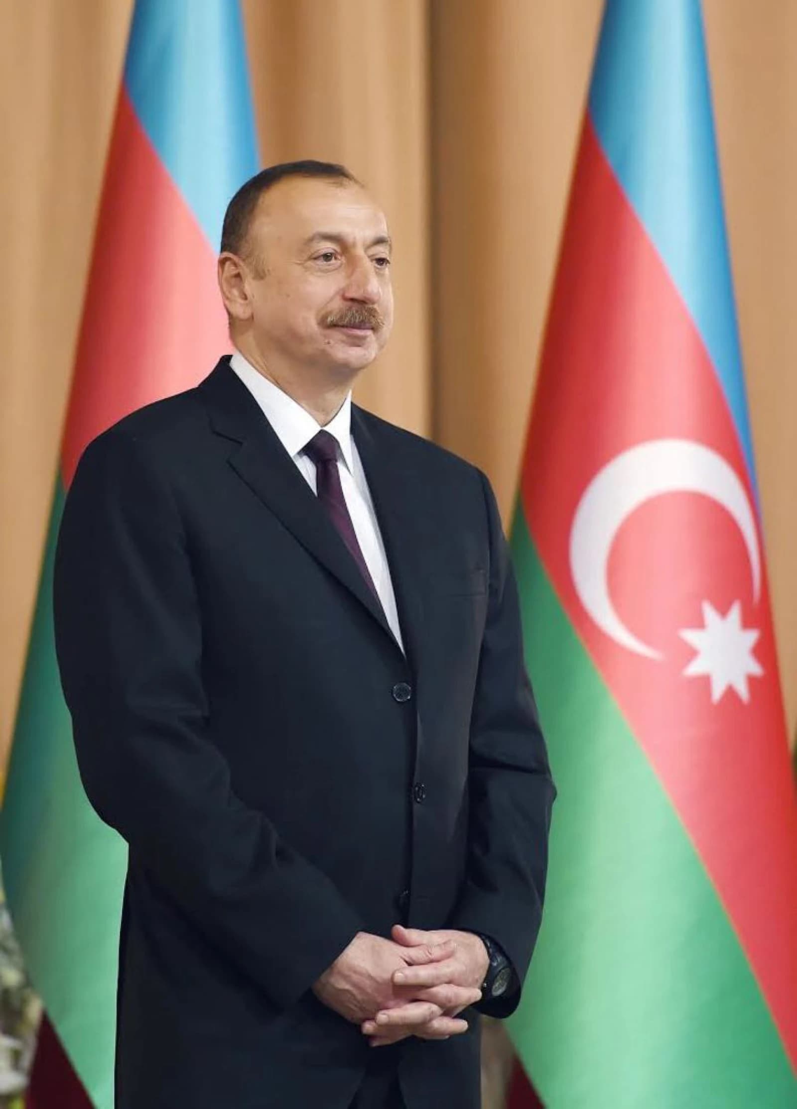 Ilham Aliyev, President of Azerbaijan, stands with clasped hands in front of national flags.