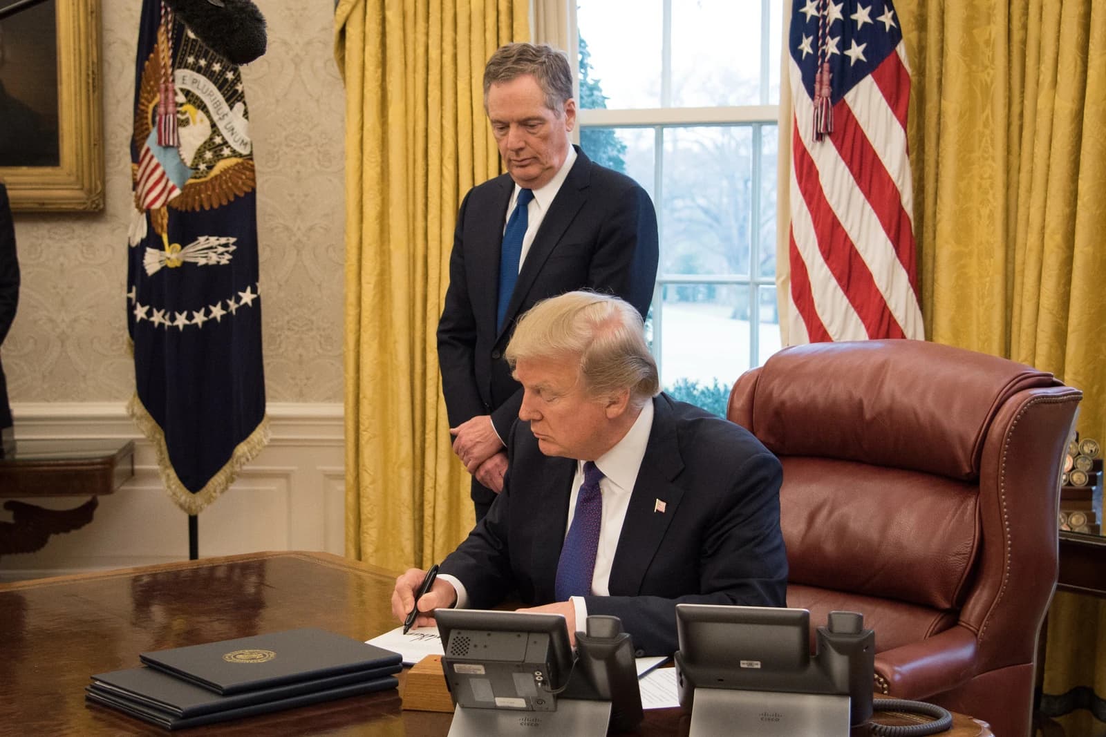 President Donald Trump signs documents at a desk as Robert Lighthizer watches.