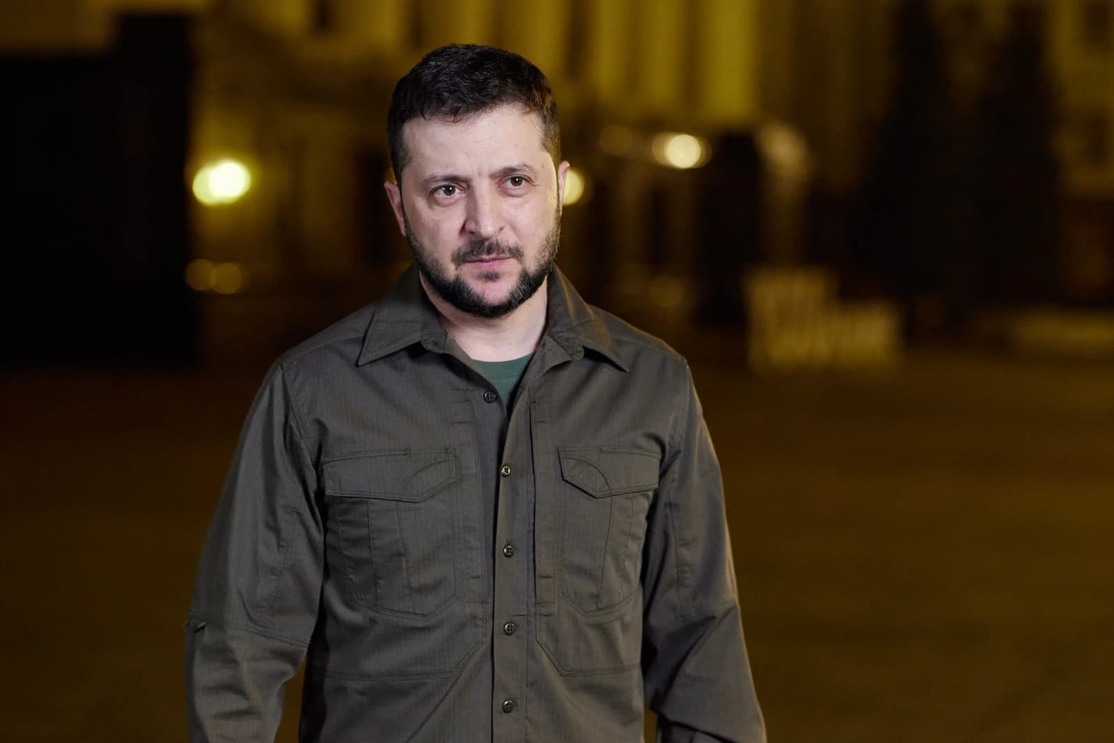 Volodymyr Zelenskyy, President of Ukraine, stands looking forward in a dark setting.