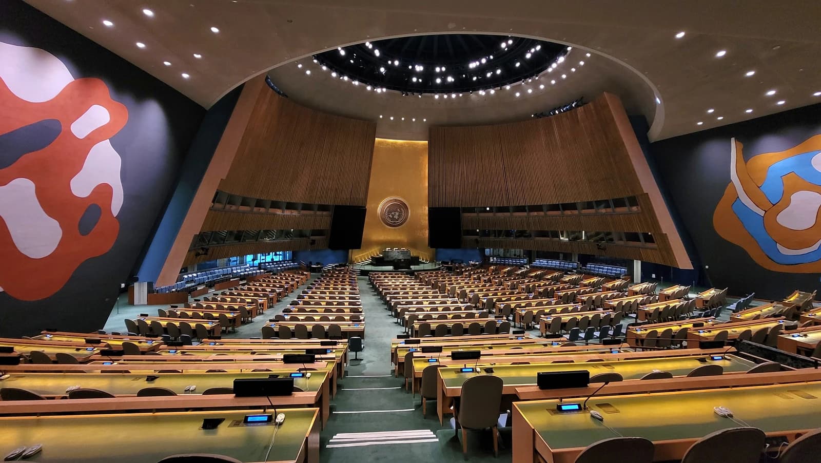 Empty United Nations General Assembly Hall with rows of desks and the UN emblem.