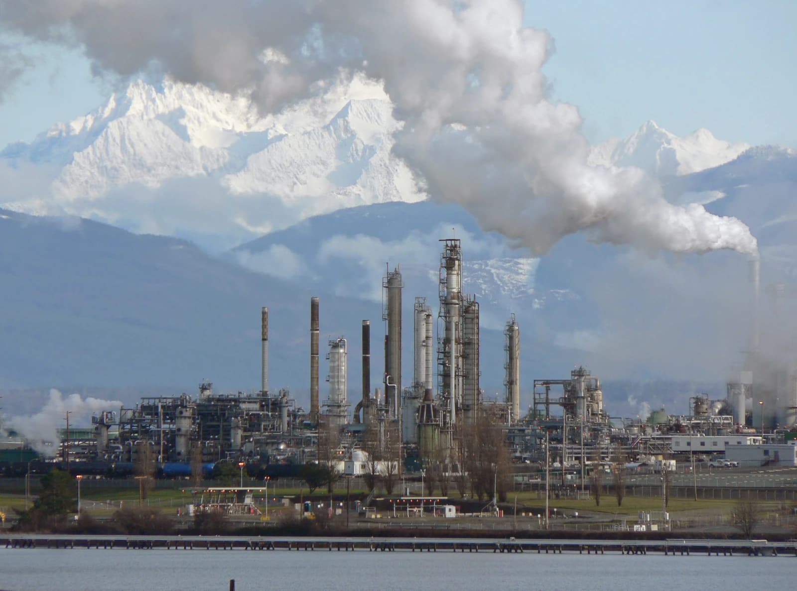 Anacortes Refinery with smoke rising from industrial structures and snow-capped mountains behind.