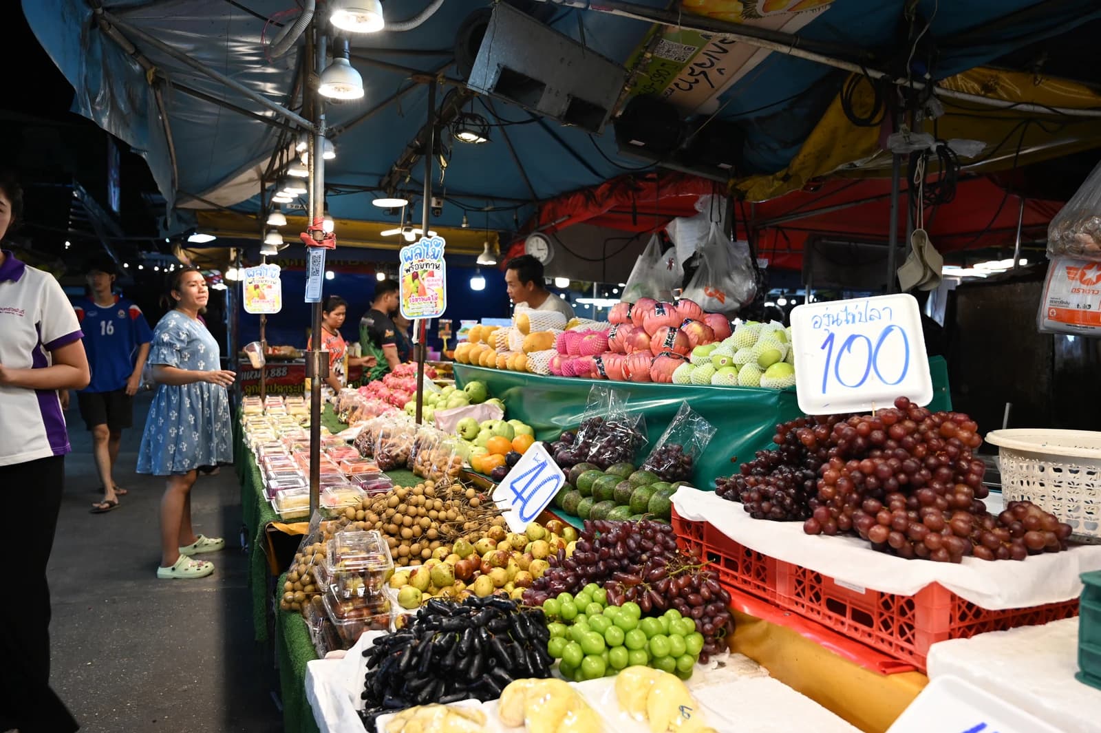 Busy night market stall overflowing with fresh fruits under bright hanging lights.