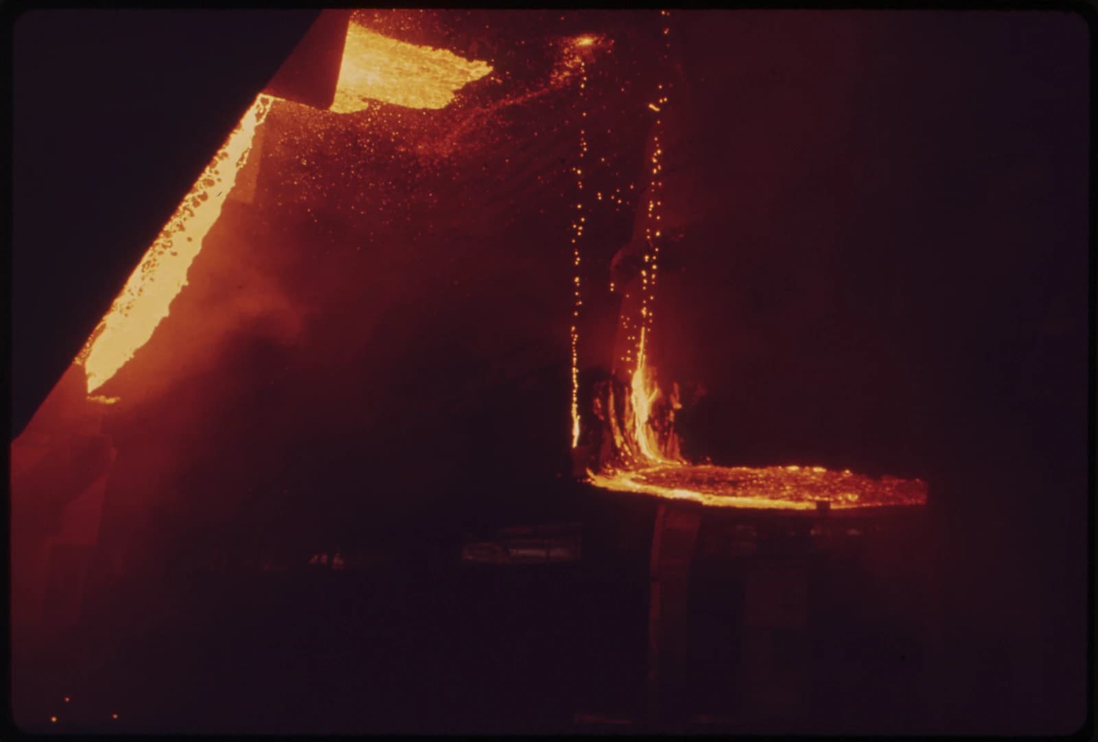 Molten steel glowing orange and yellow pours into a channel inside a dark factory.