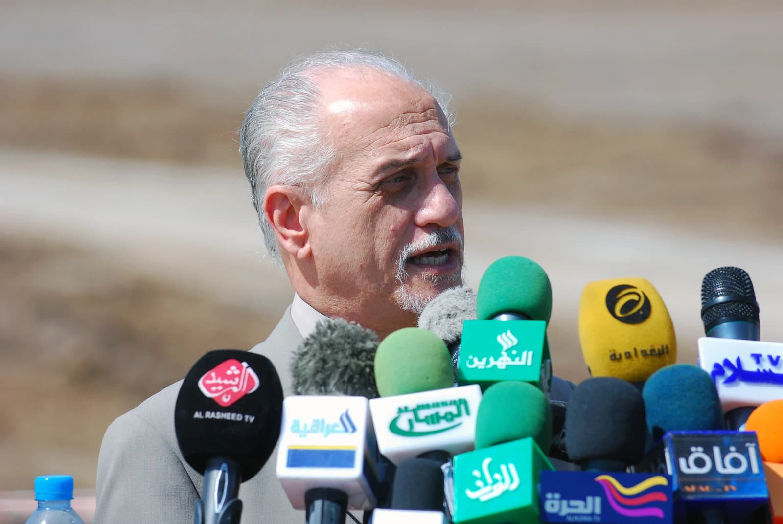An Iraqi official speaks at a podium surrounded by microphones from various news organizations.