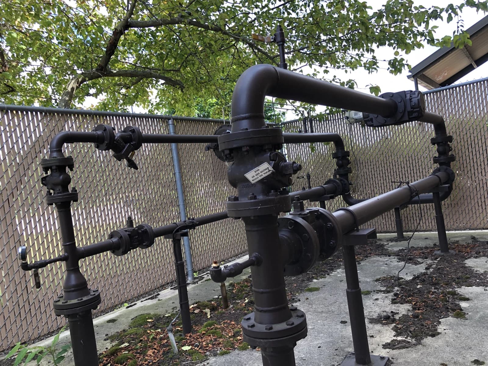 Natural gas pipeline station with dark pipes and valves visible against a chain link fence.