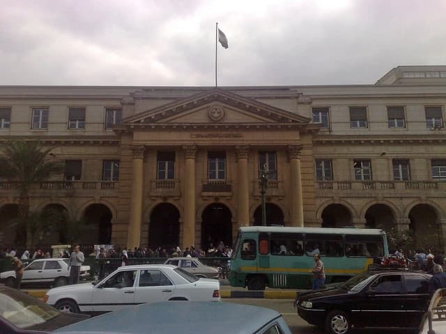 Exterior view of a government building with an Egyptian flag and traffic in the foreground.