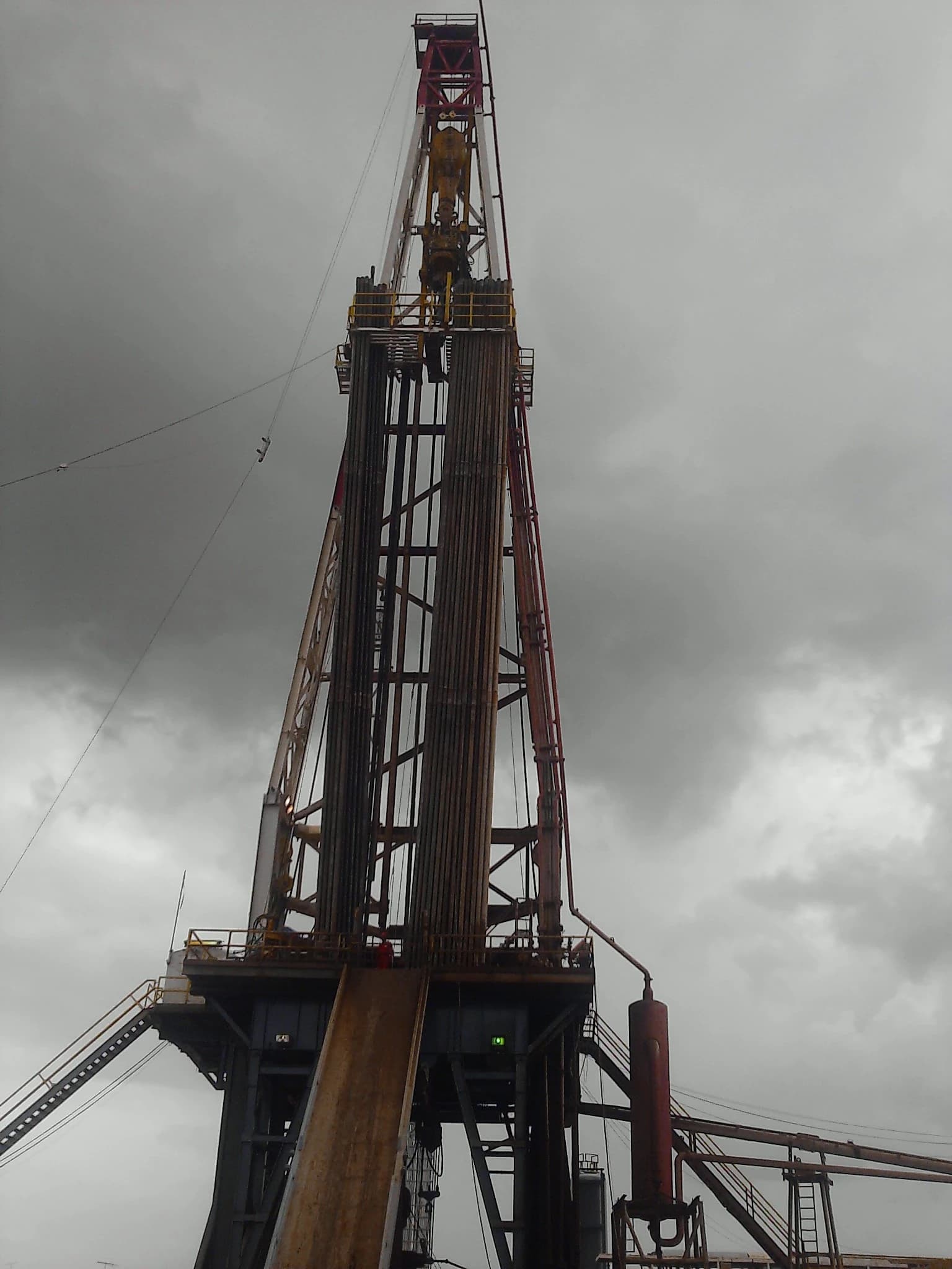 An oil drilling rig tower with pipes and platforms against a grey sky.