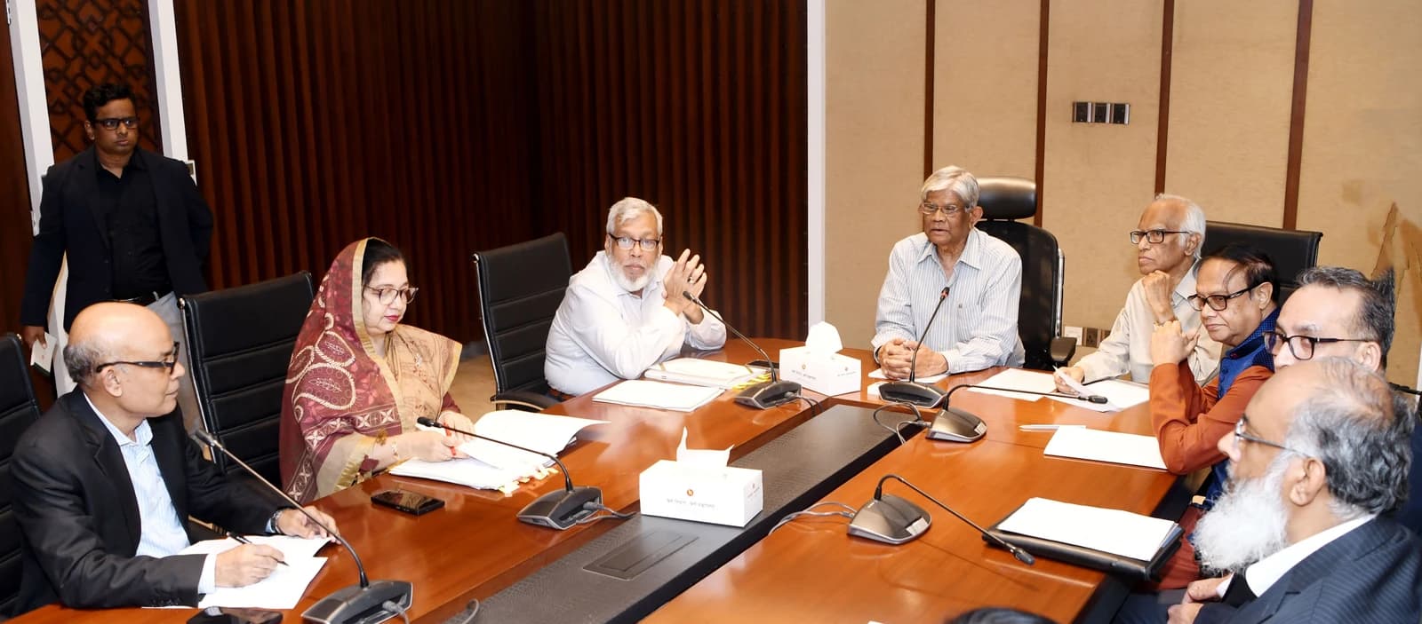 Meeting participants, including a woman in a headscarf, seated at a conference table in Dhaka.