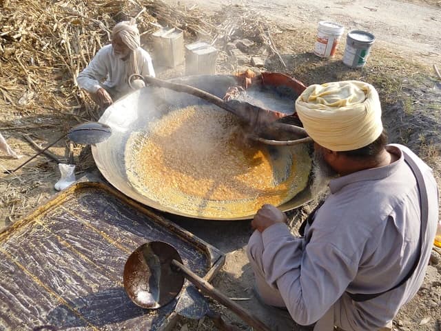 Two men cooking jaggery in a large, shallow pan outdoors.