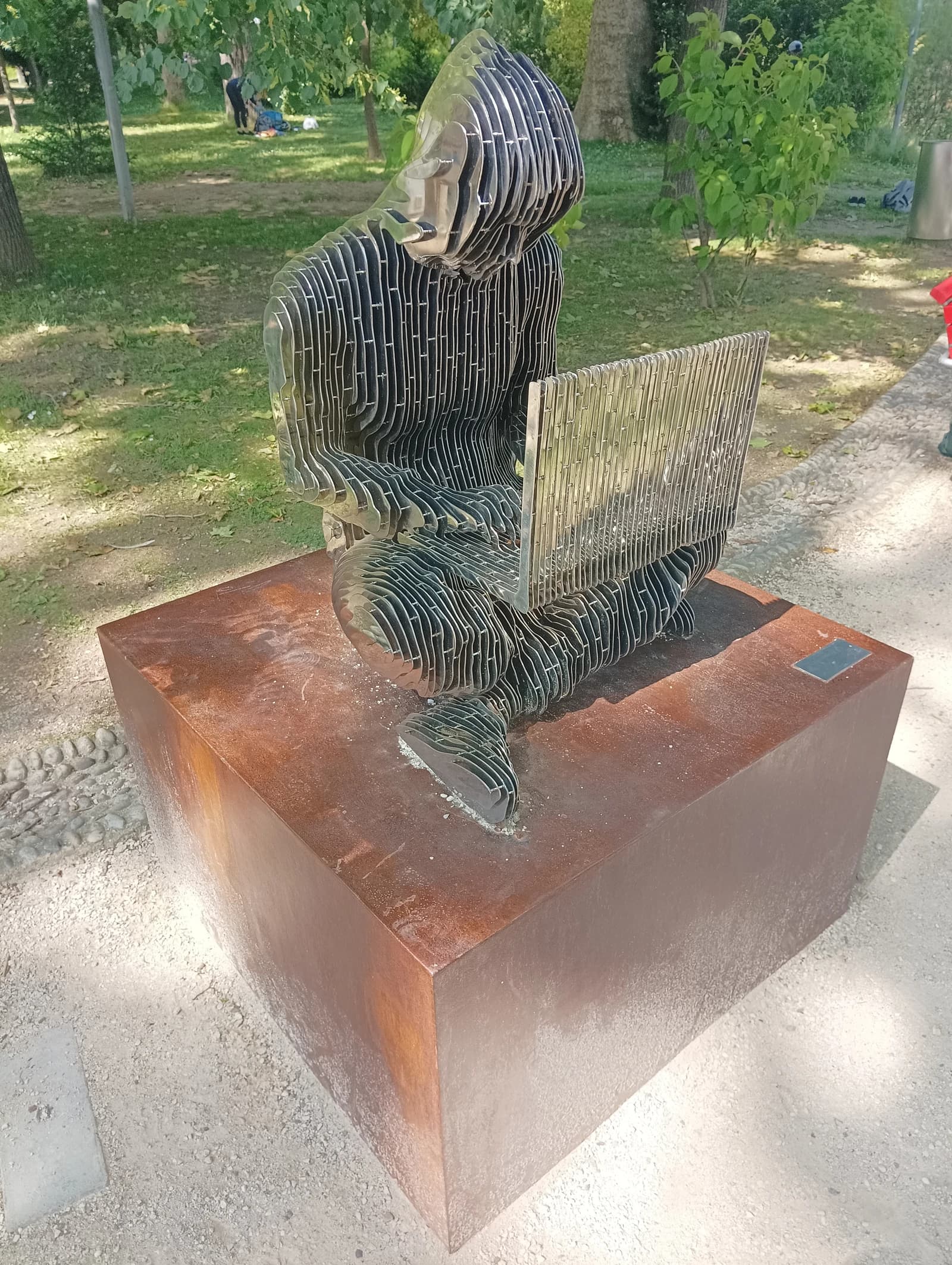 A metallic statue of Satoshi Nakamoto seated with a laptop in a park setting.