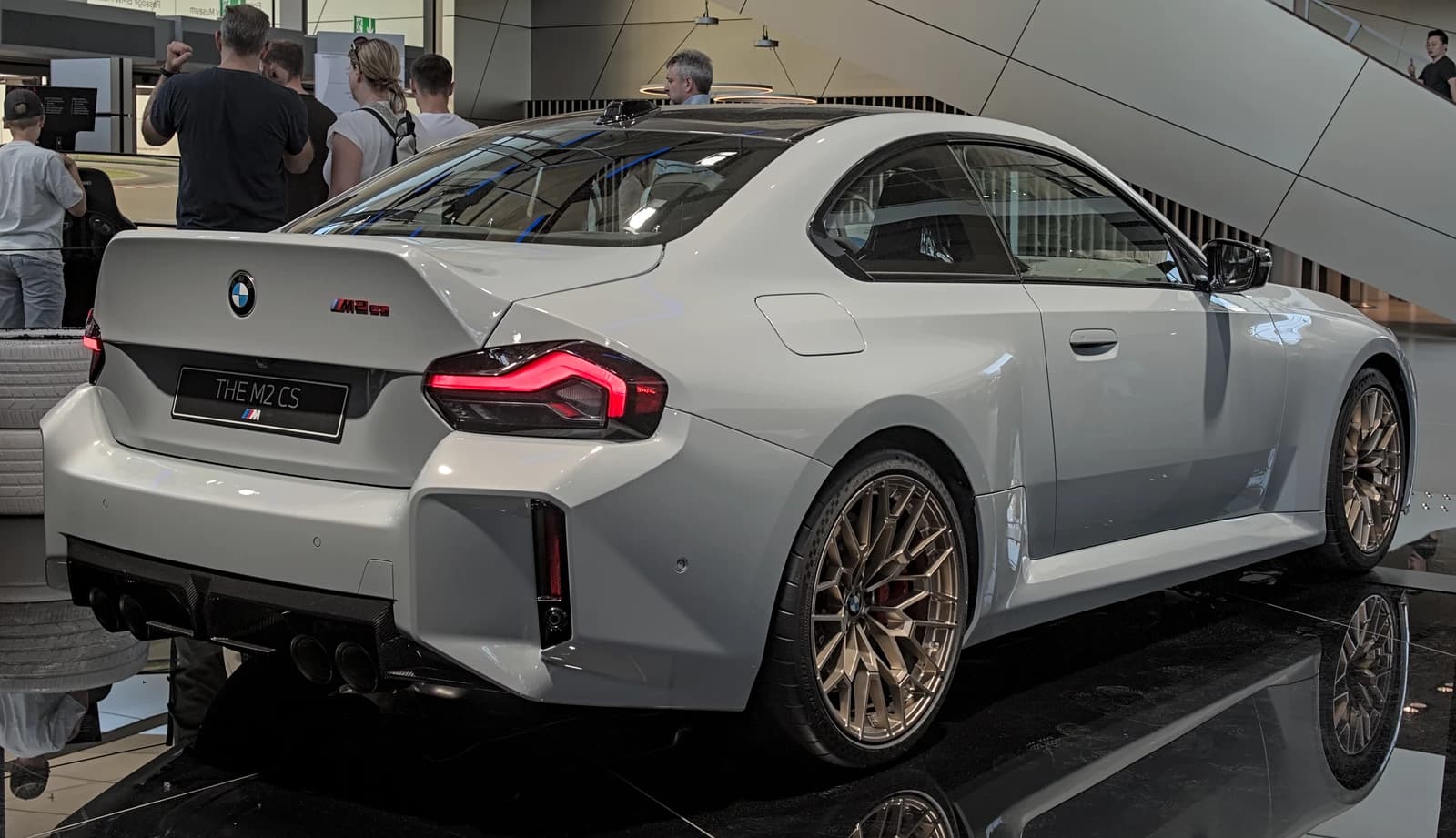 Light gray BMW M2 CS car with golden wheels displayed indoors, viewed from the rear.