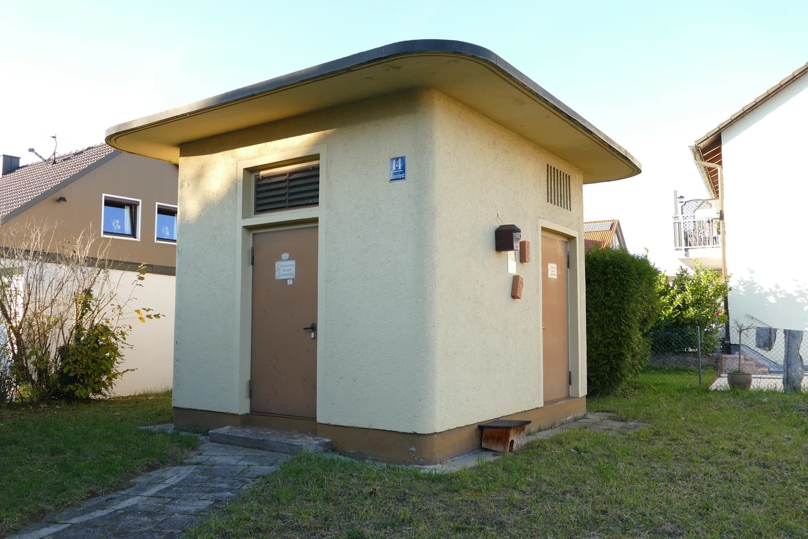 Small, light-colored transformer station with two doors, surrounded by grass and residential houses.