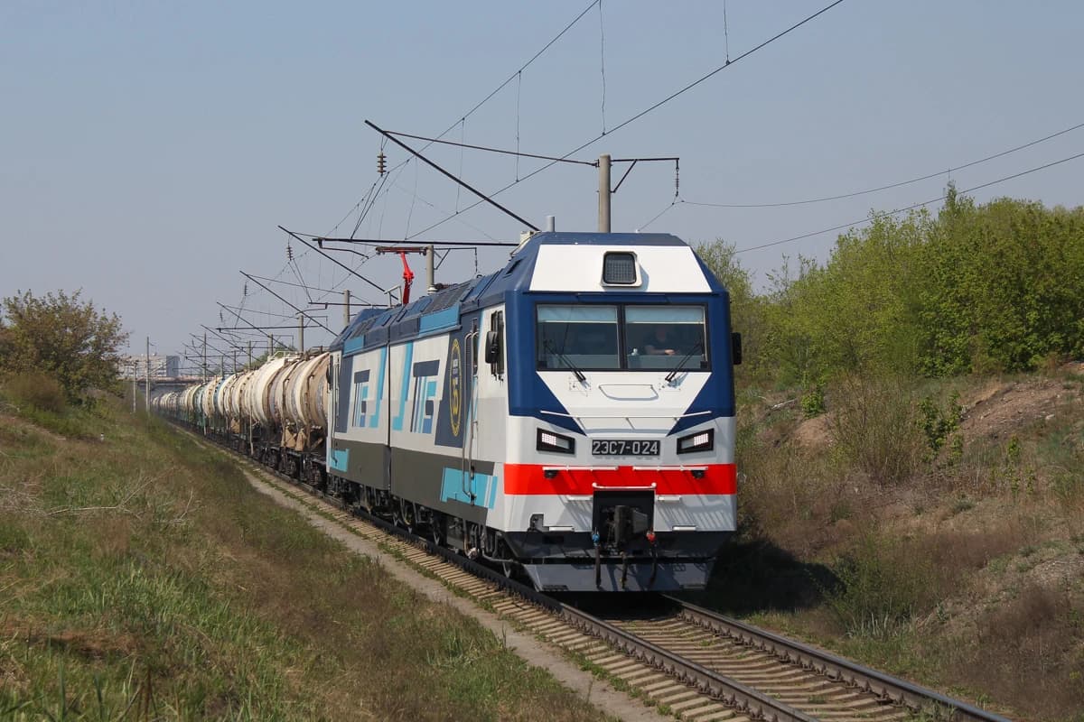 2ES7-024 electric locomotive pulling a long train of tanker wagons through a rural landscape.