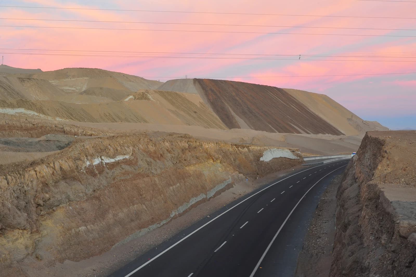 An empty highway winds through a desert landscape with large mining waste piles under a colorful sky.