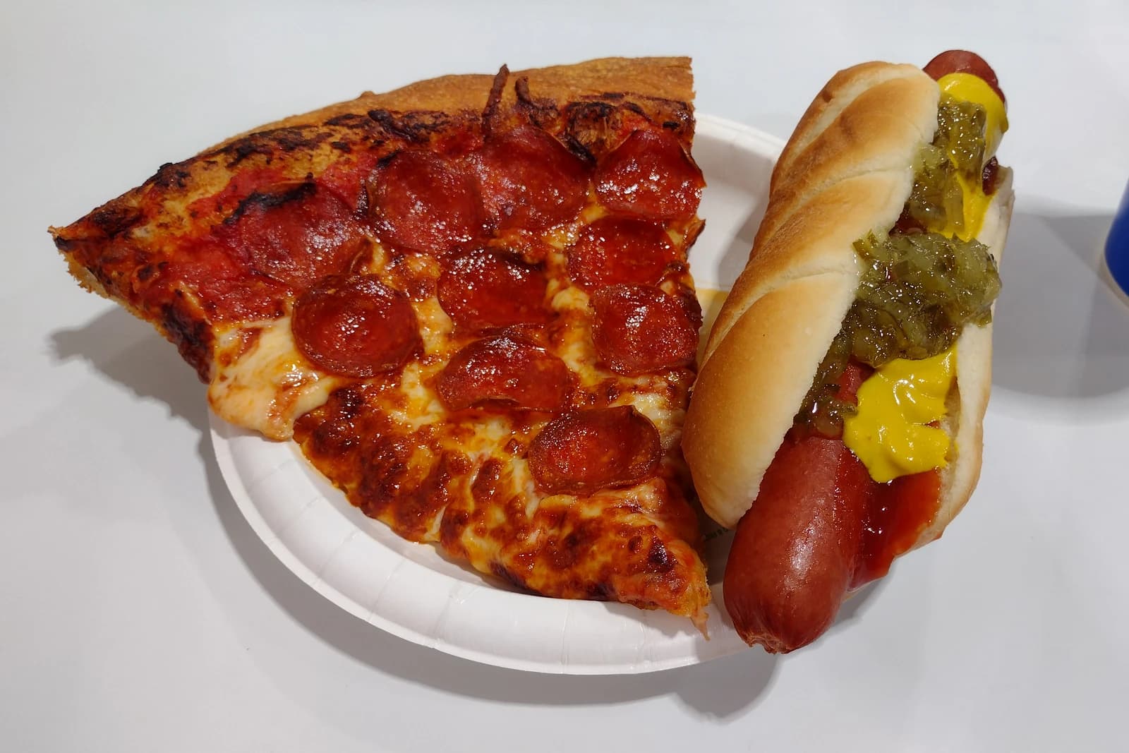A pepperoni pizza slice and a hot dog with mustard and relish on a white paper plate.