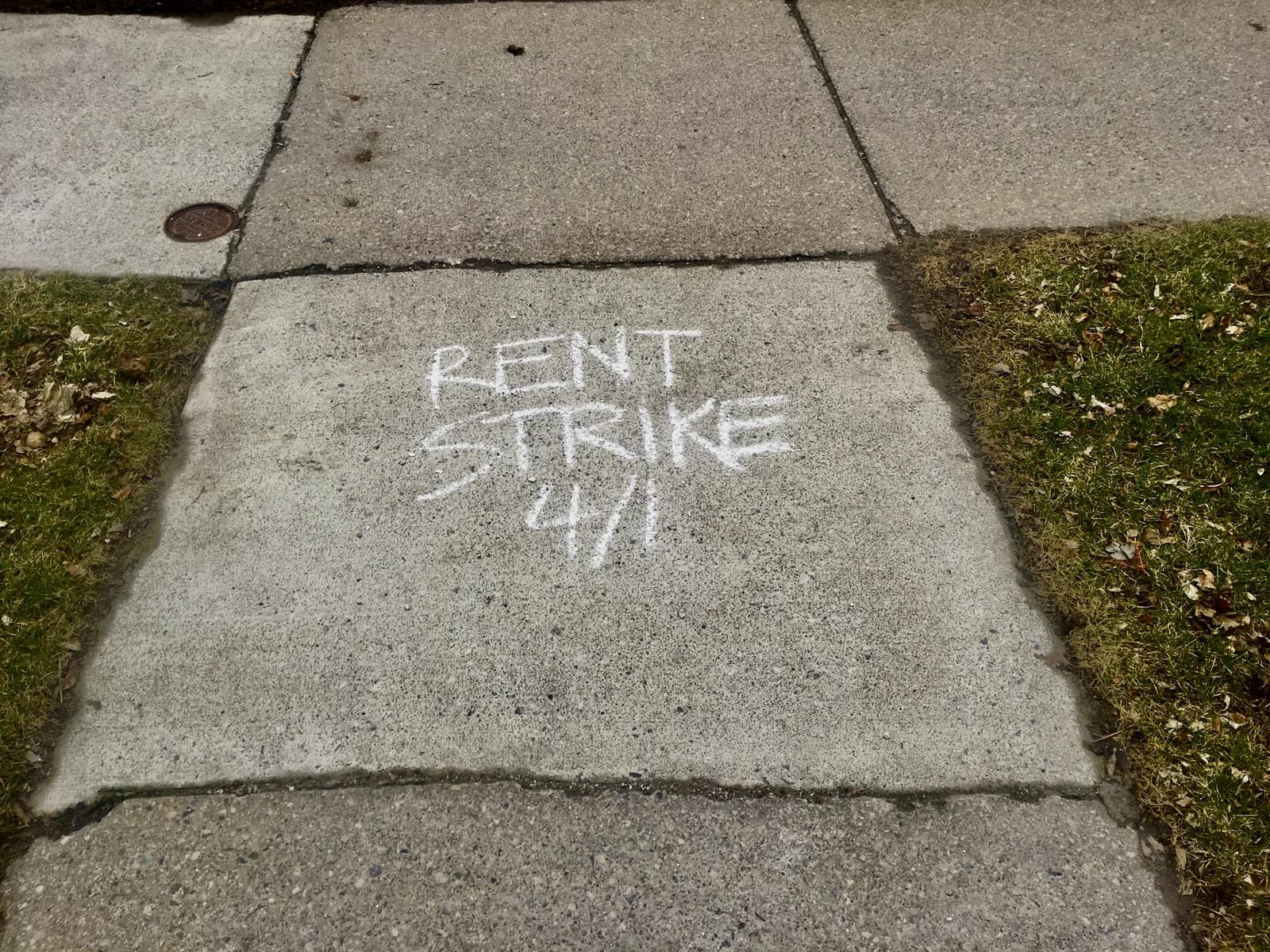 Chalk graffiti on a sidewalk reads "Rent Strike 4/1" next to green grass.