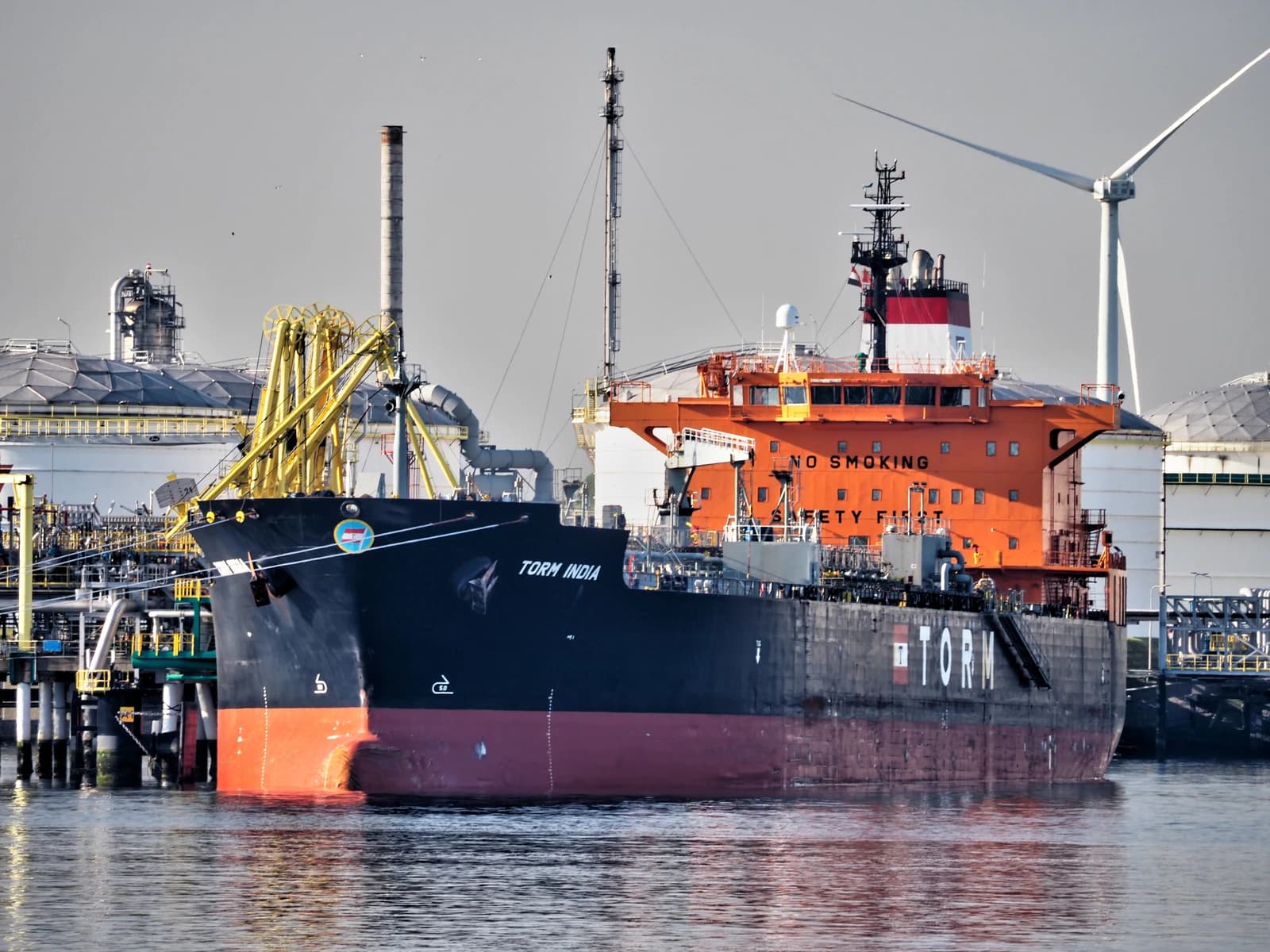 Oil-chemical tanker TORM INDIA docked at a port next to industrial storage tanks and a wind turbine.