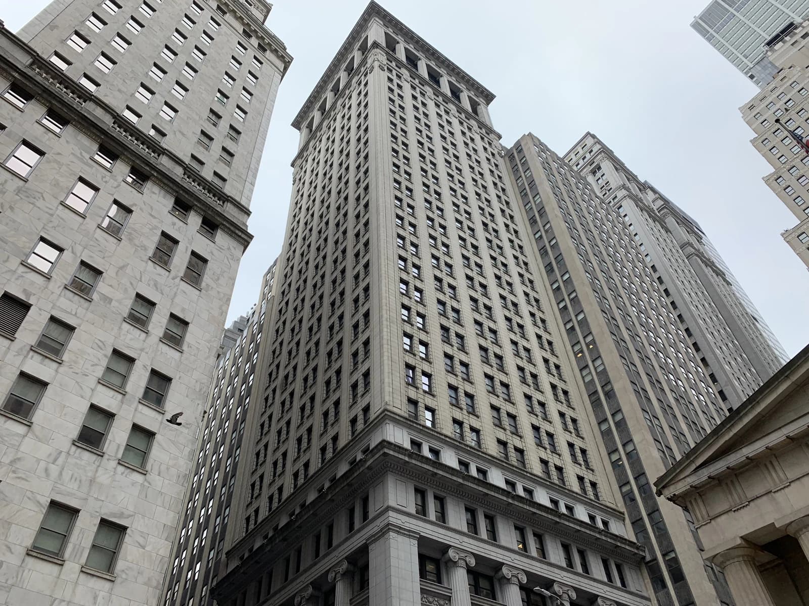 Tall, grey Wall Street buildings with many windows, viewed from below on a cloudy day.