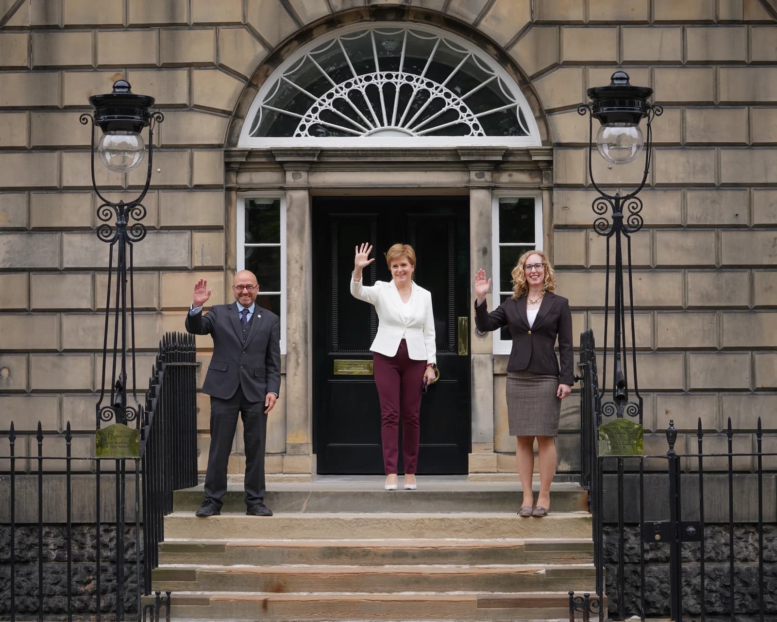 Nicola Sturgeon, First Minister of Scotland, with Scottish Green co-leaders Patrick Harvie and Lorna Slater, waving from a doorway.