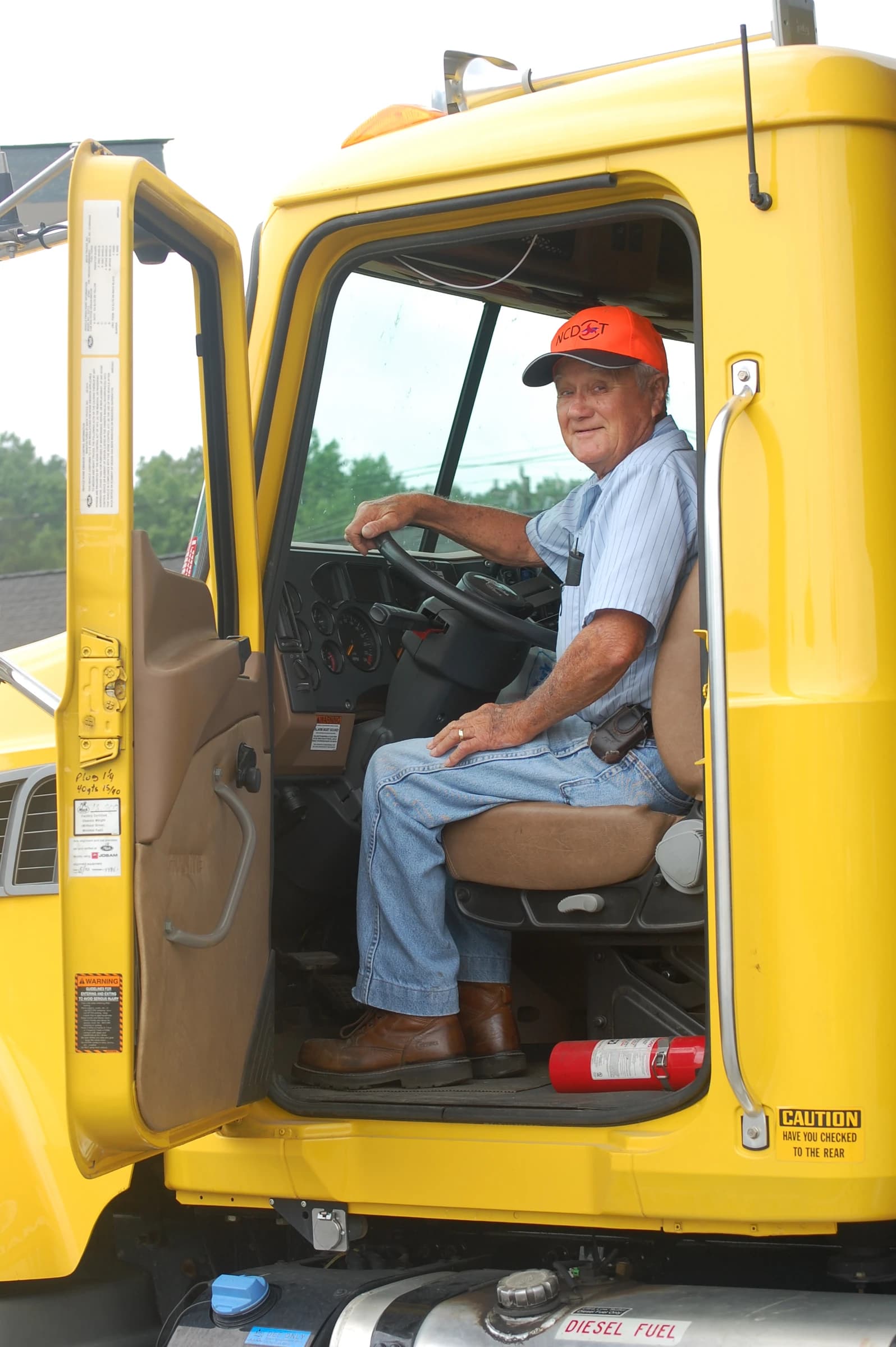 Man wearing an orange hat and striped shirt sits in the driver's seat of a yellow dump truck.
