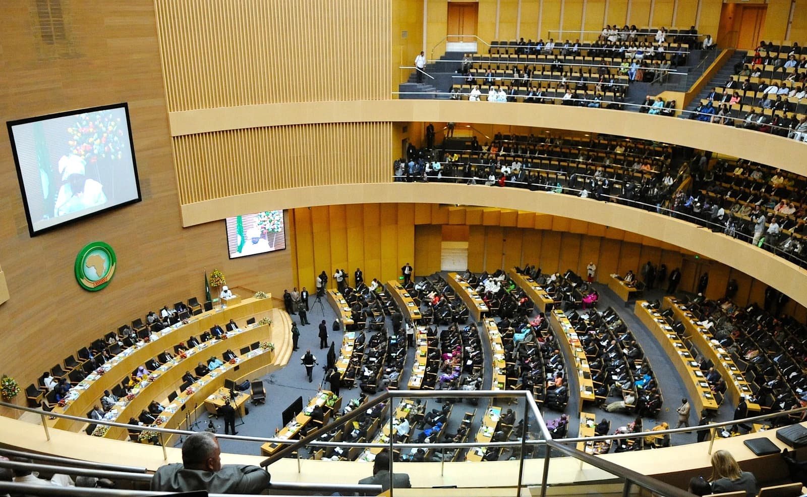 People seated in a large, tiered hall with a screen displaying a speaker.