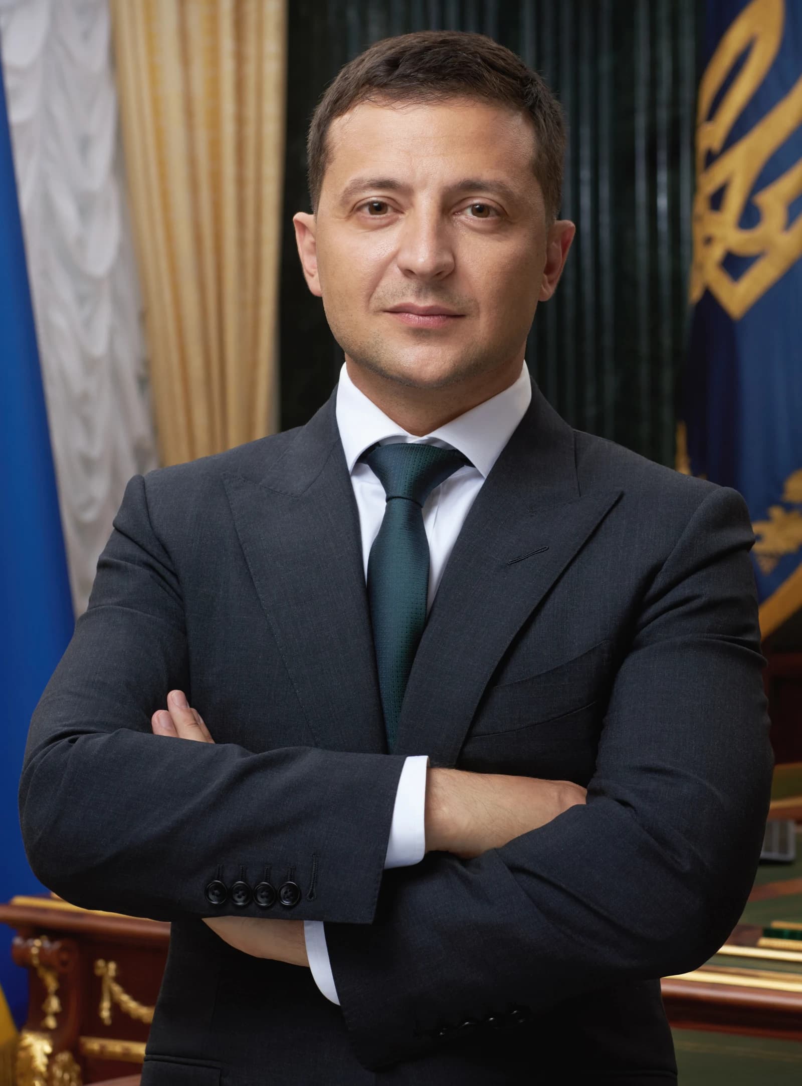 Volodymyr Zelensky, President of Ukraine, stands with arms crossed in an official portrait.