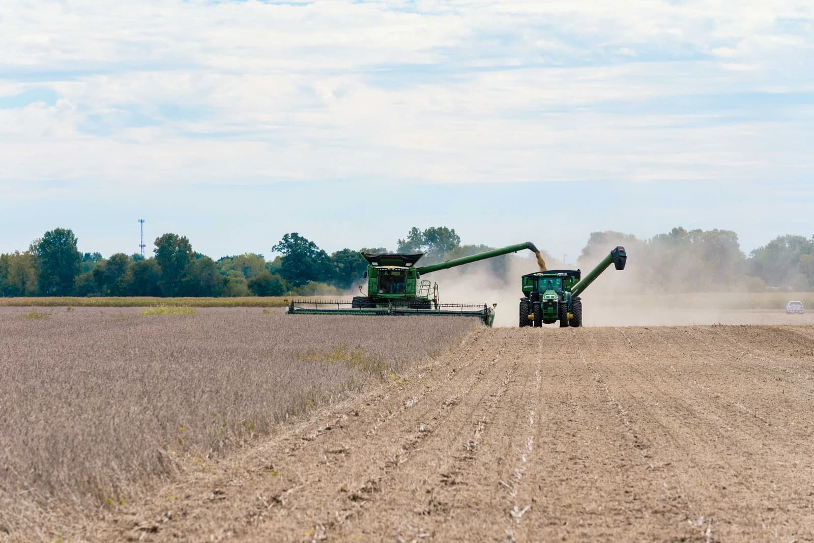 Combine harvester emptying harvested soybeans into a tractor's grain cart in a field.