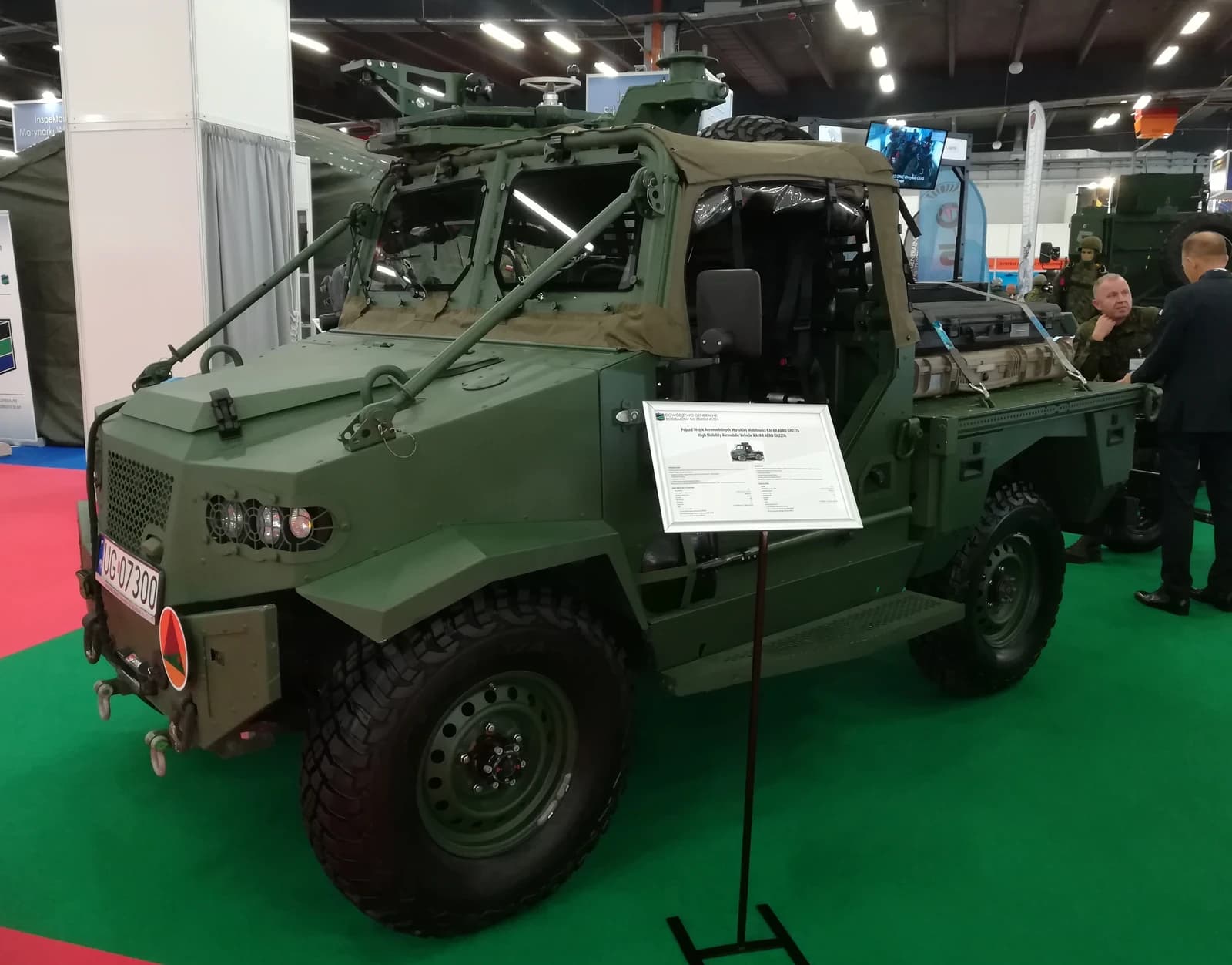 Military vehicle displayed indoors at an exhibition hall, with two men standing nearby.