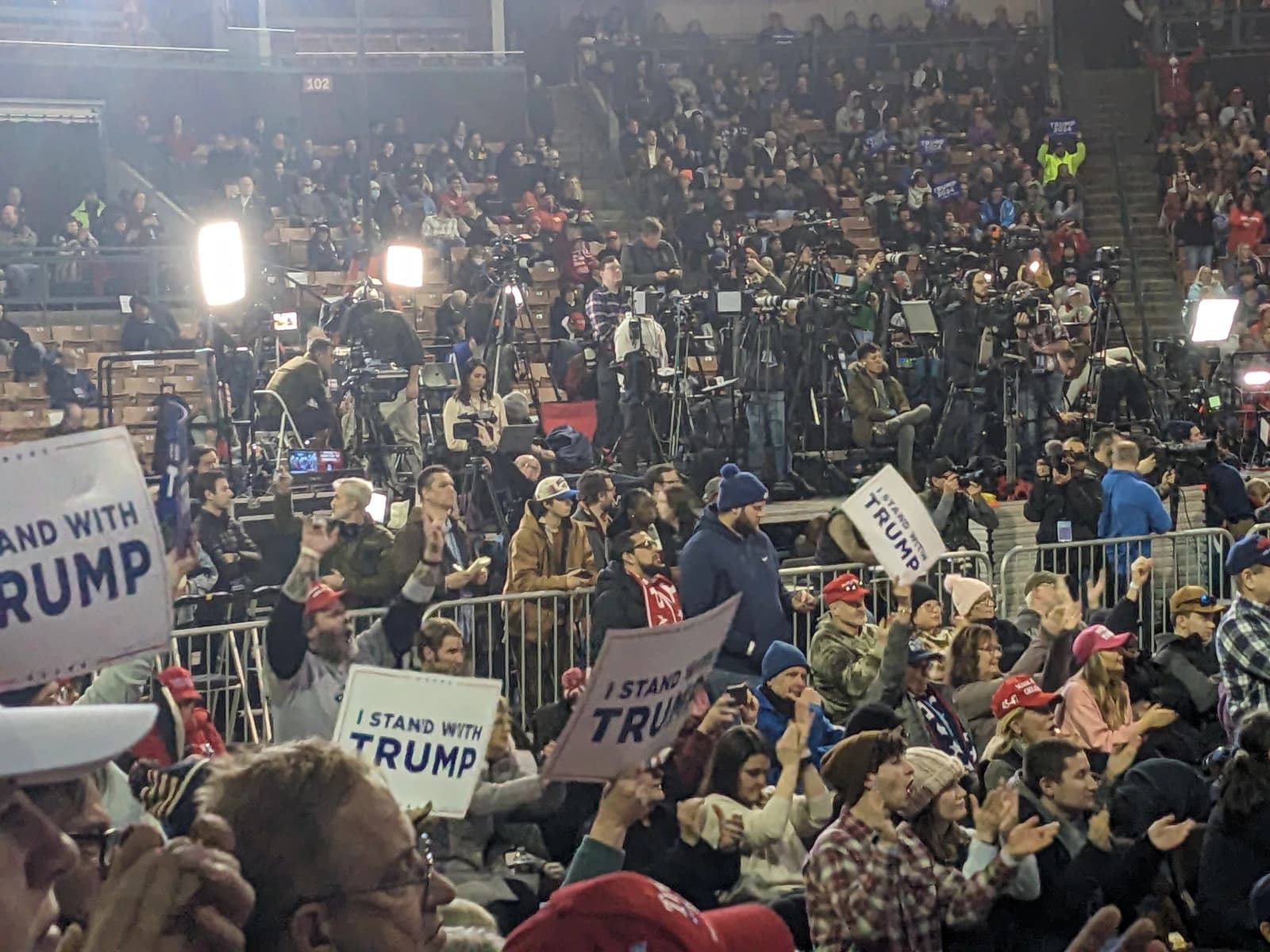 An audience holds 'I Stand with Trump' signs at a rally with media cameras in the background.