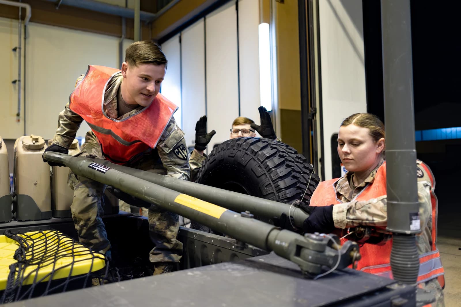 Military personnel in camouflage uniforms and orange vests lifting equipment during training.