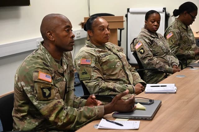 Several soldiers in camouflage uniforms seated at a conference table with notebooks and laptops.