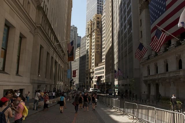 Pedestrians walk along Broad Street in Manhattan, New York, with tall buildings visible.