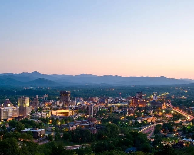 City of Asheville, North Carolina at dusk, with buildings lit up and mountains in the distance.