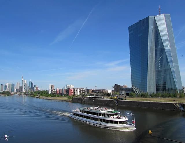 A large modern glass skyscraper, the European Central Bank building, stands by a river with a boat.