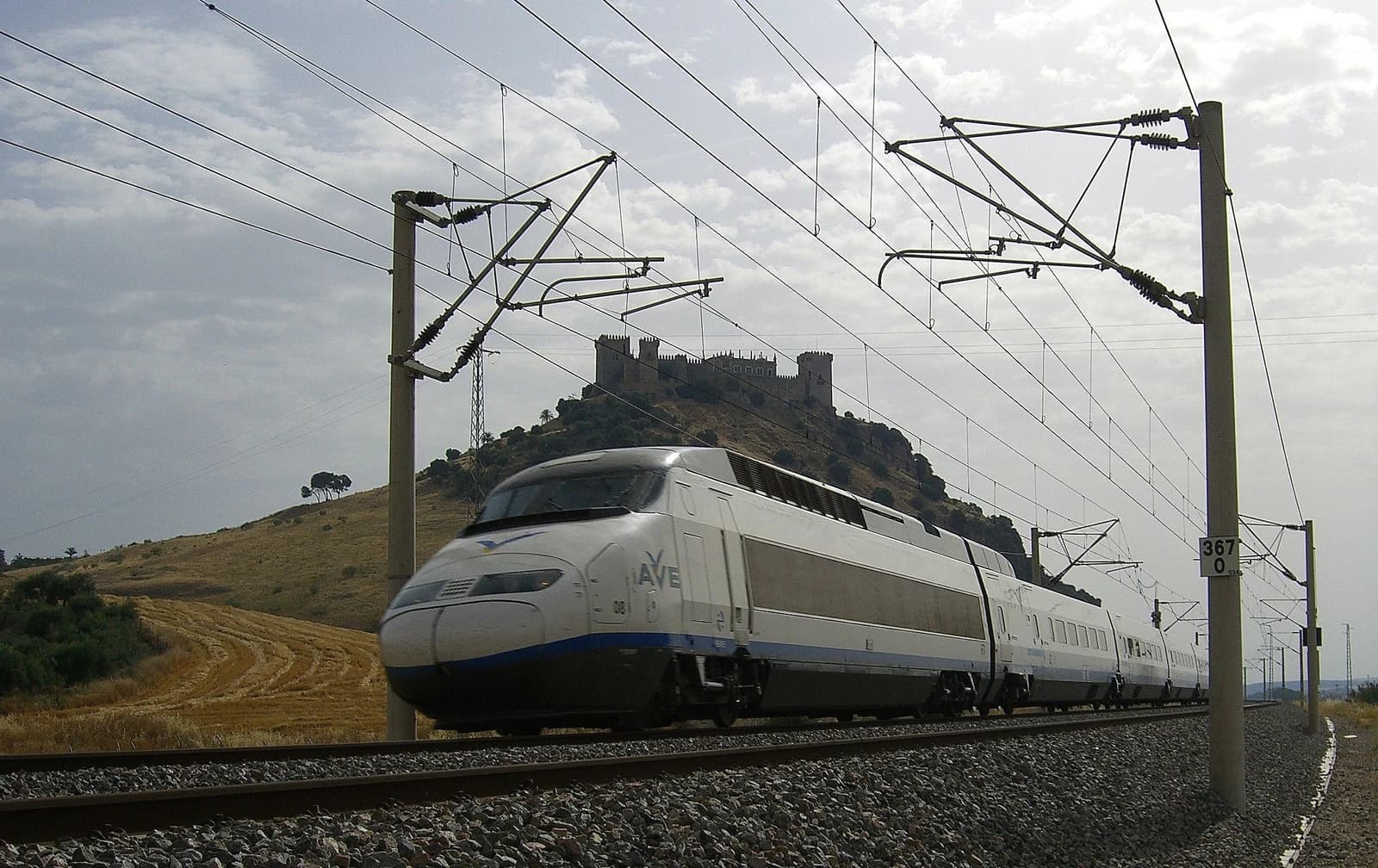 An AVE Serie 100 high-speed train travels on tracks through a rural landscape.