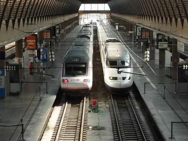 Two high-speed trains are parked side-by-side on tracks inside Santa Justa Station in Seville, Spain.