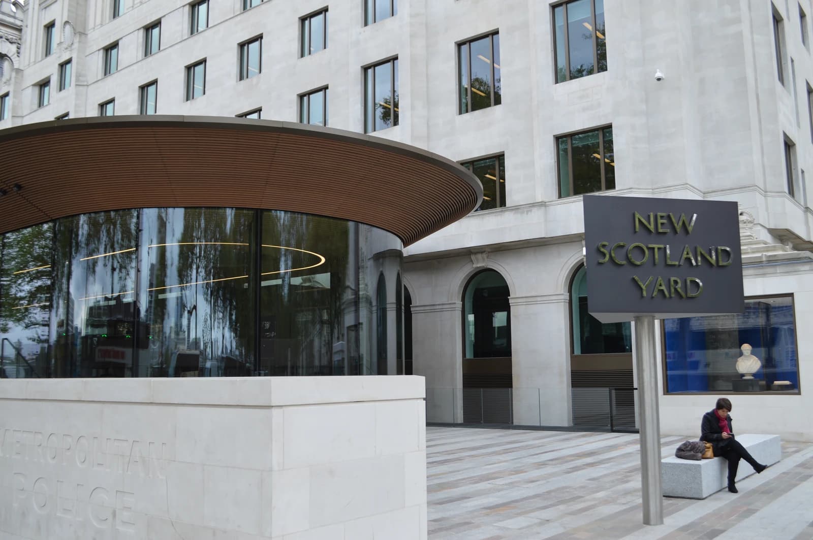 Exterior of New Scotland Yard building with its sign, a woman sits on a bench.
