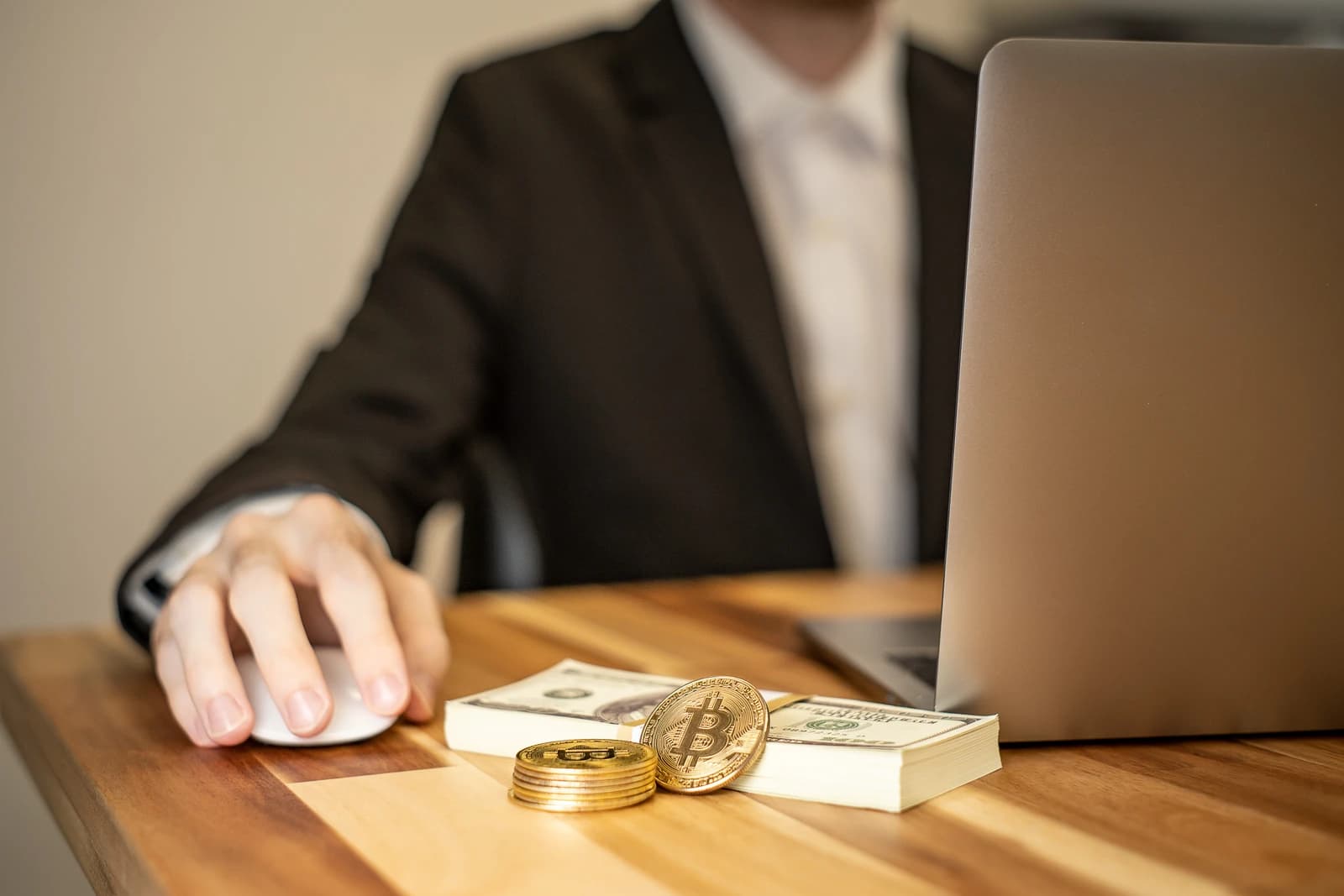 Person in a suit uses computer mouse with cryptocurrency and cash on a desk.