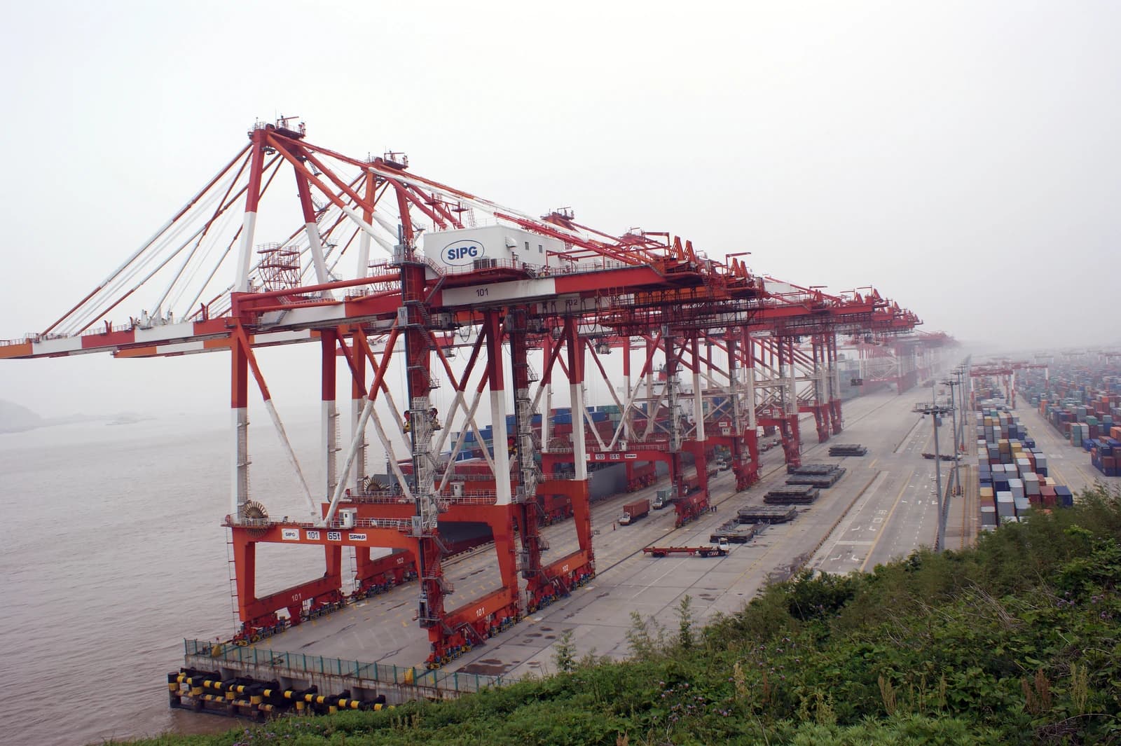 Red and white container cranes stand along the water's edge at Shanghai Yangshan Port.