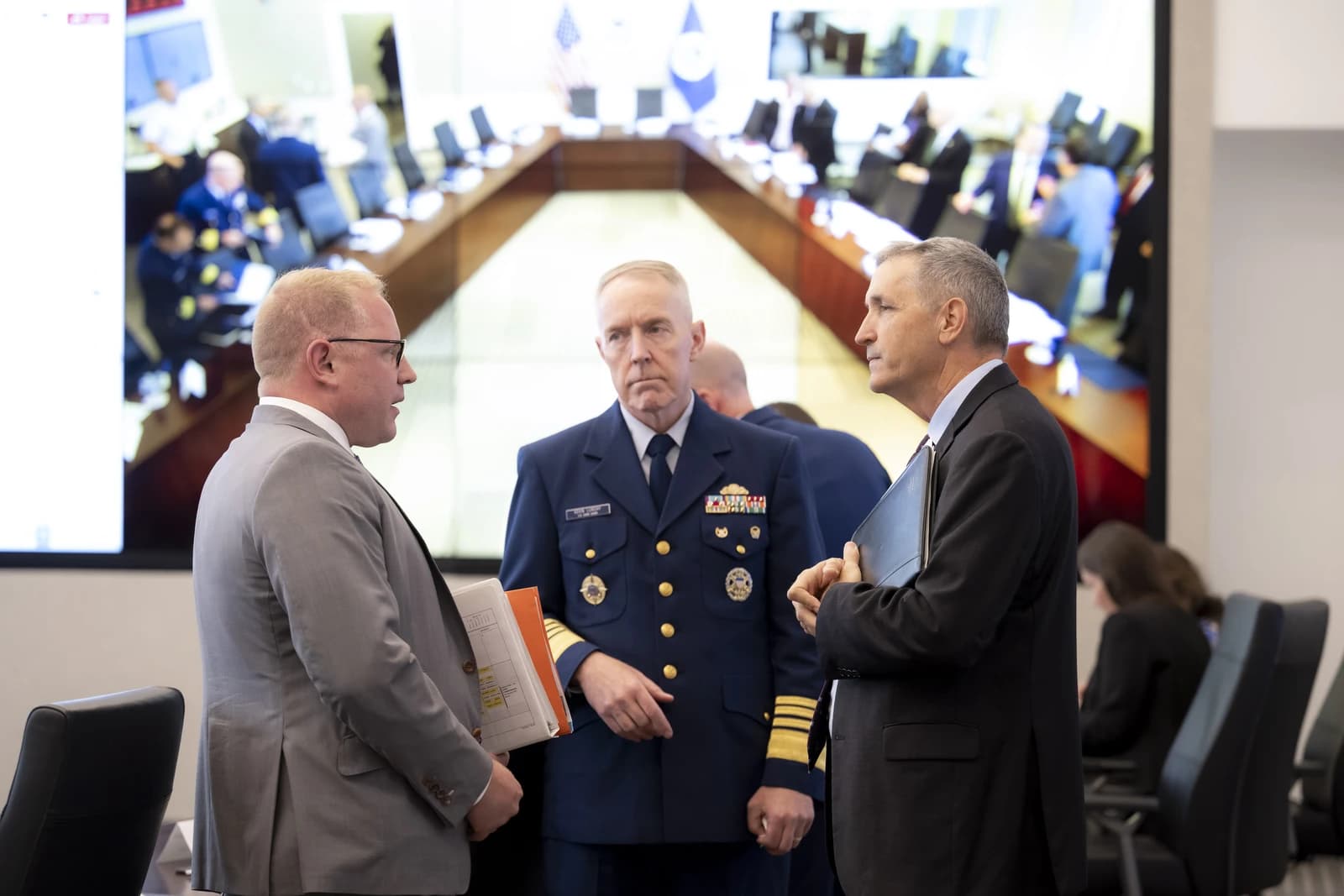 Three men in suits and uniform speaking in a meeting room.