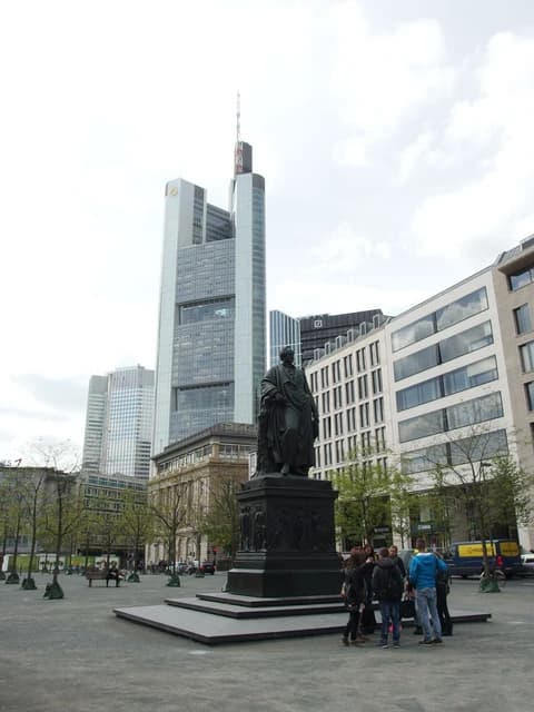 A statue of a figure in a robe stands in a square with modern high-rise buildings in the background.