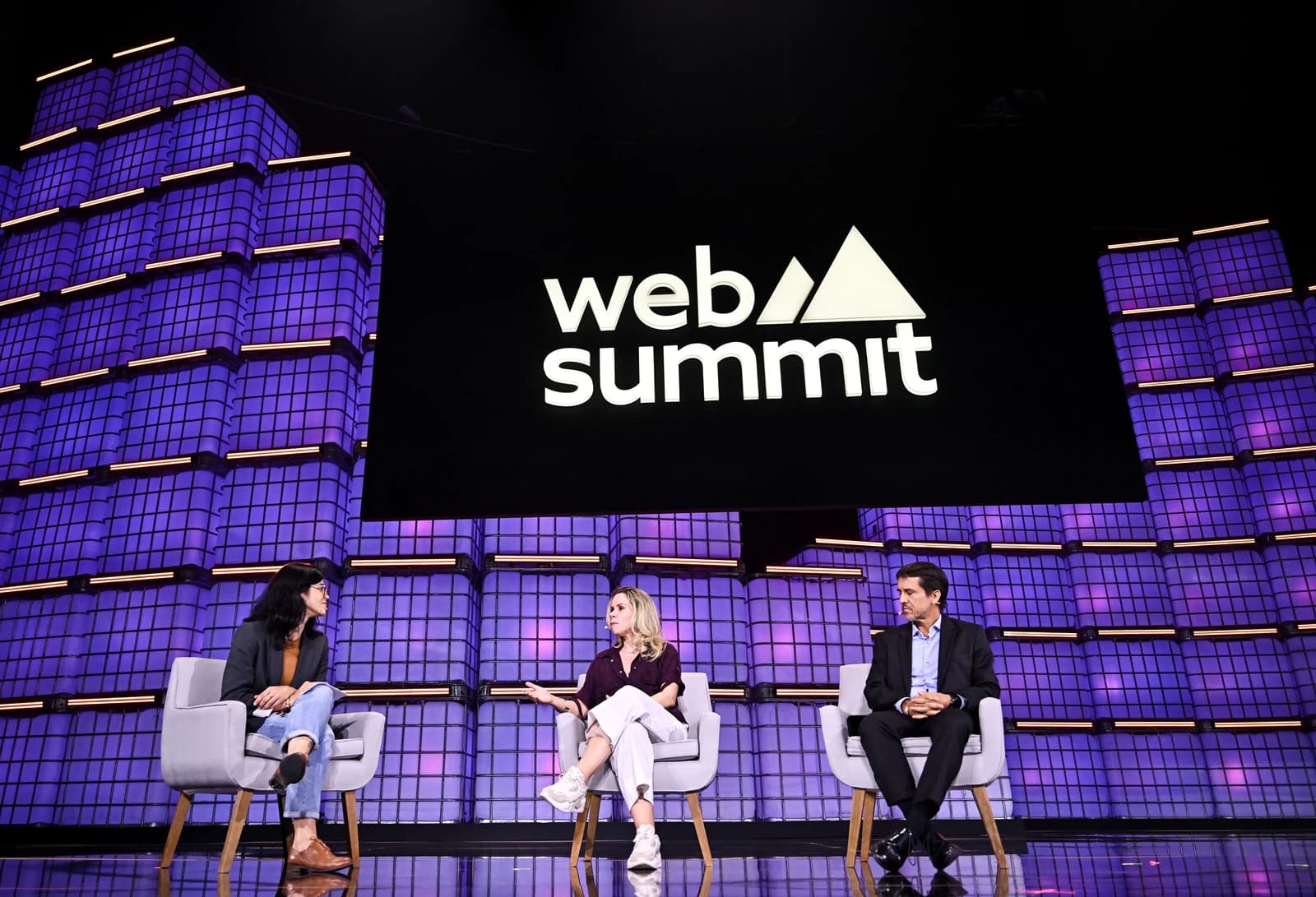 Three individuals seated in chairs on a stage with a 'Web Summit' logo in the background.