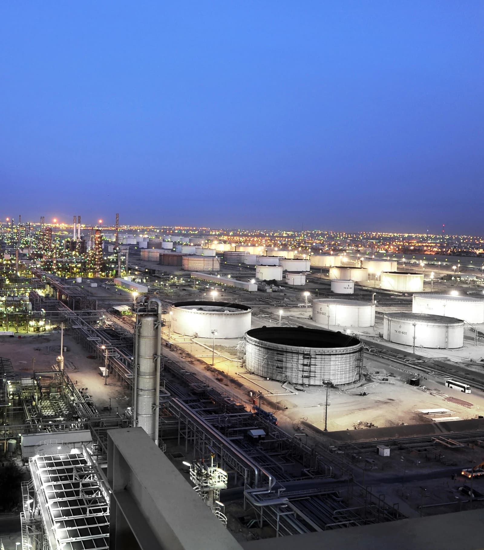 An industrial landscape of a large oil refinery with numerous illuminated storage tanks and pipes at dusk.